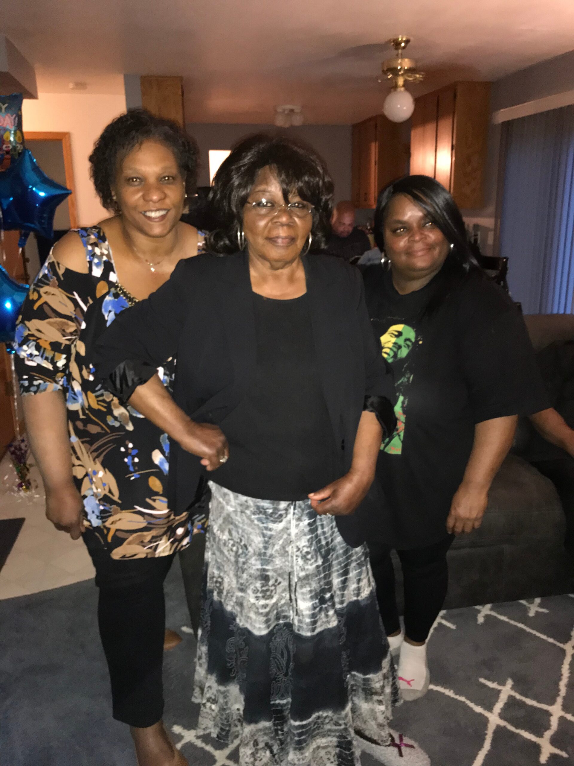 Three women posing together indoors, smiling at the camera: left in a floral dress, center in black, right in a black shirt with a graphic tee.