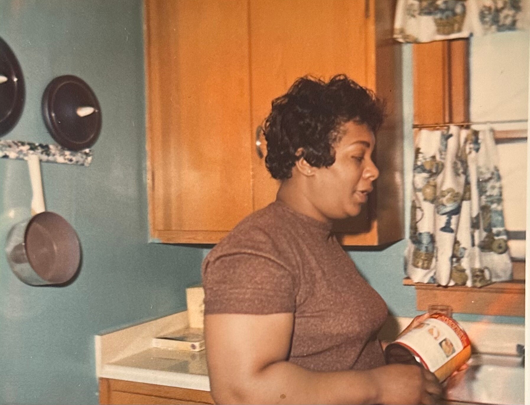 Woman in a brown shirt stands at a kitchen counter, opening a cylindrical can in a mid‑century kitchen.