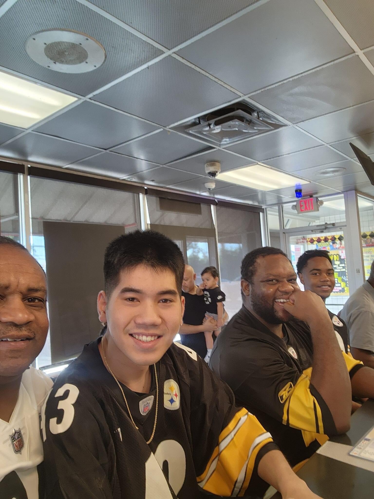 Group of men in team jerseys posing and smiling indoors, near a counter and ceiling tiles in the background