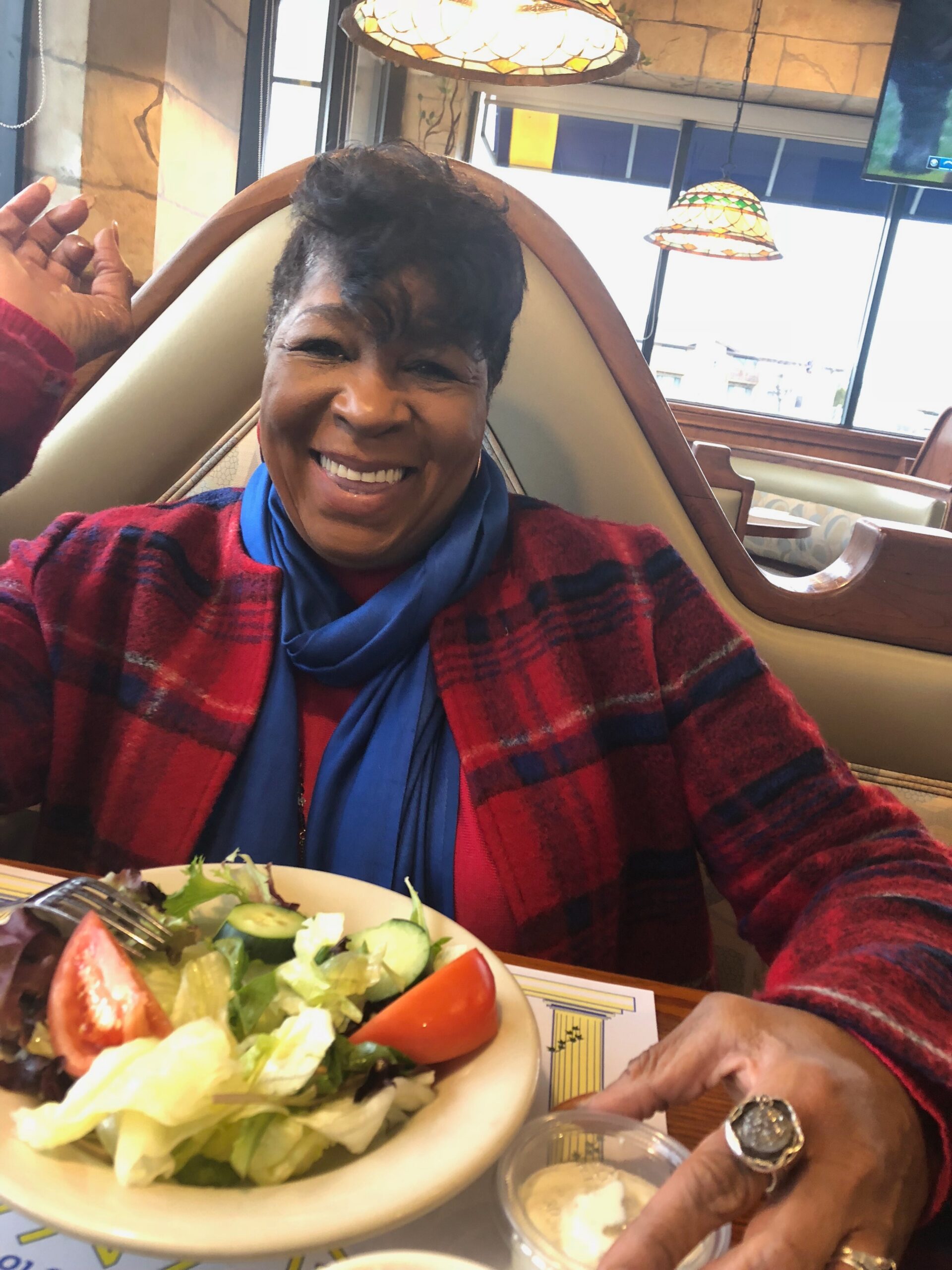 Smiling woman wearing a red plaid jacket and blue scarf sits at a restaurant table with a large salad in the foreground.