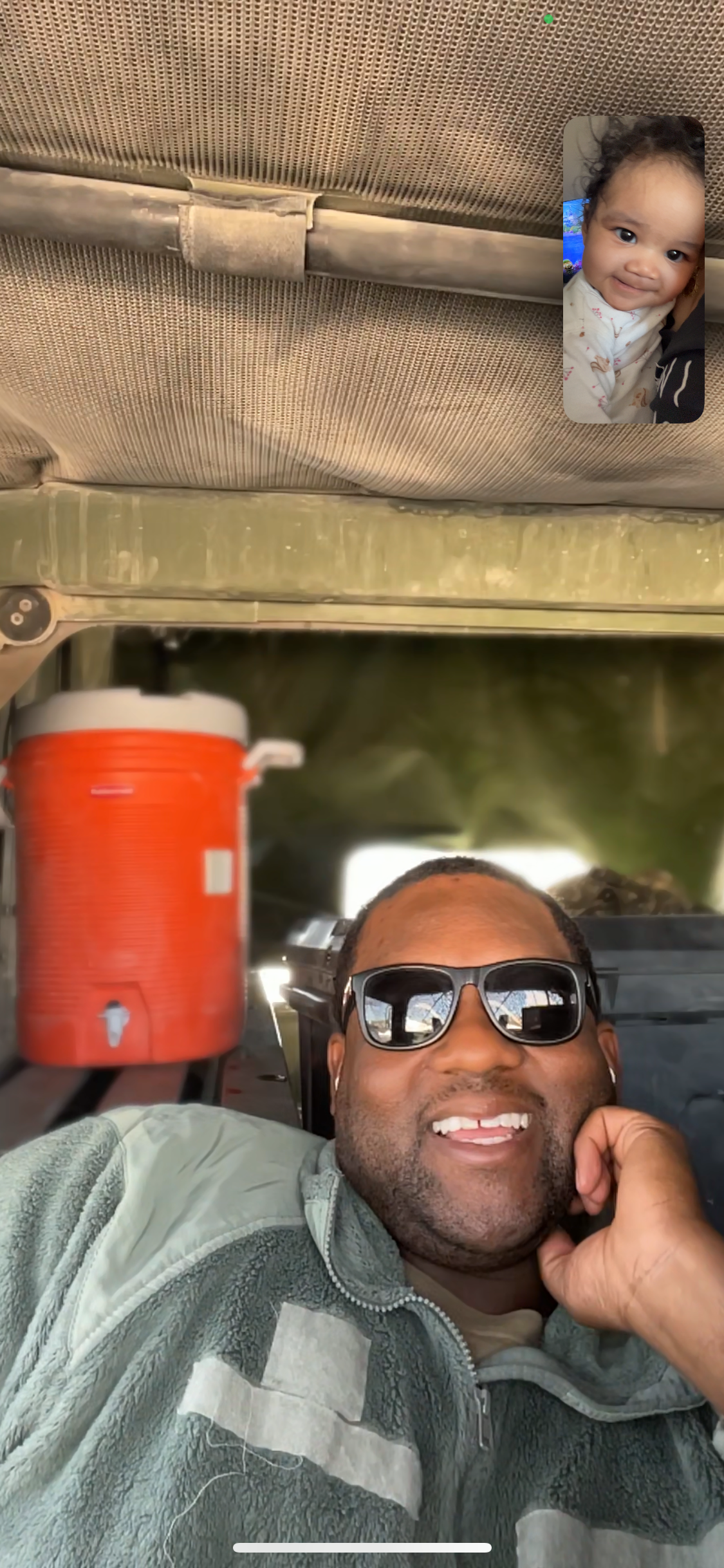 Smiling man wearing sunglasses takes a selfie inside a vehicle, with a small video call showing a baby in the top-right corner and an orange cooler in the back.