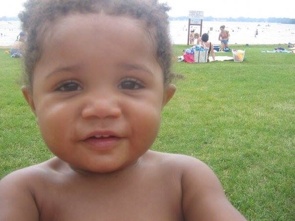 Close-up of a smiling toddler at a grassy park by the beach, with people and towels in the background.