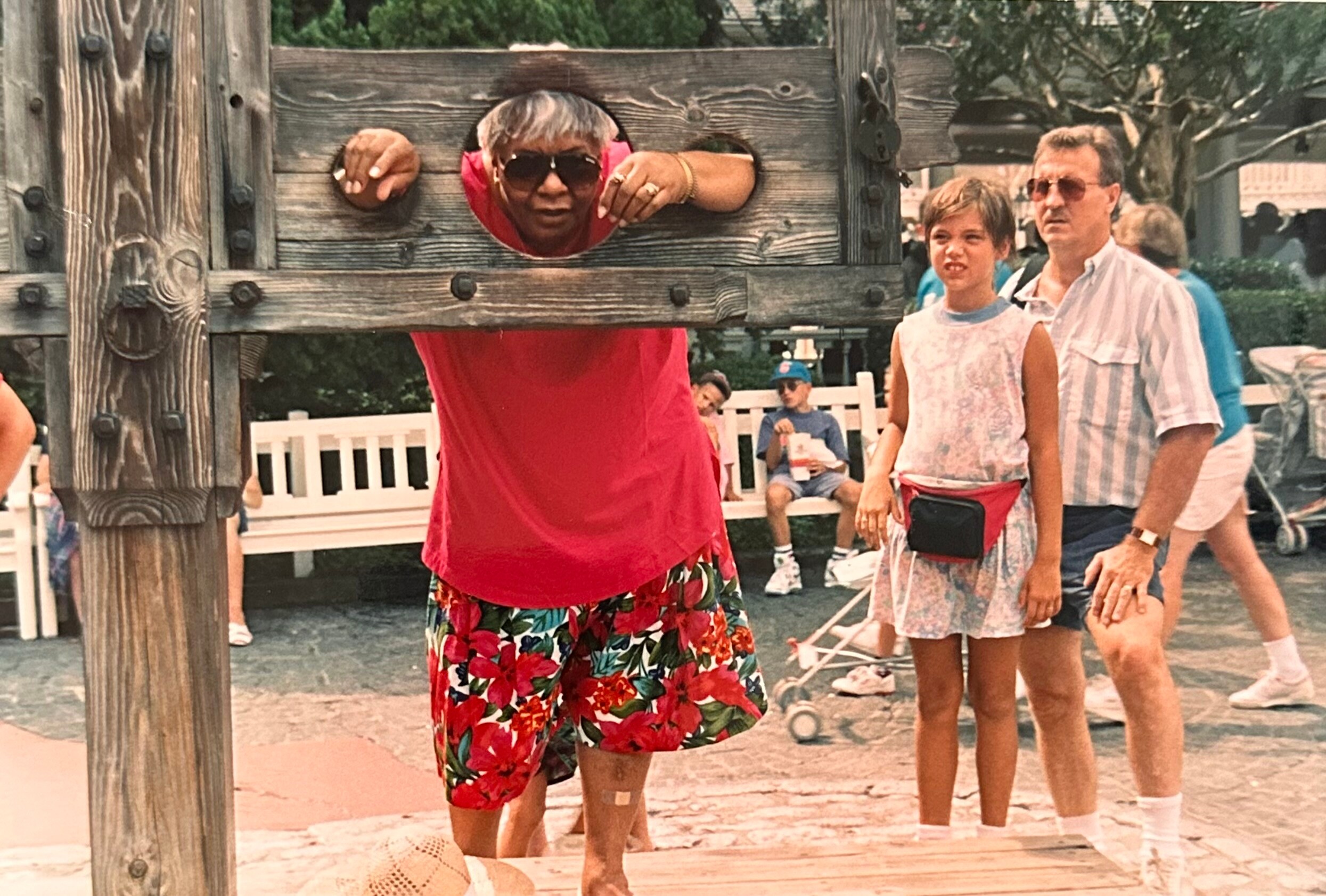 Person in a red shirt leans through a round hole in a rustic wooden frame while bystanders watch at an outdoor amusement area.