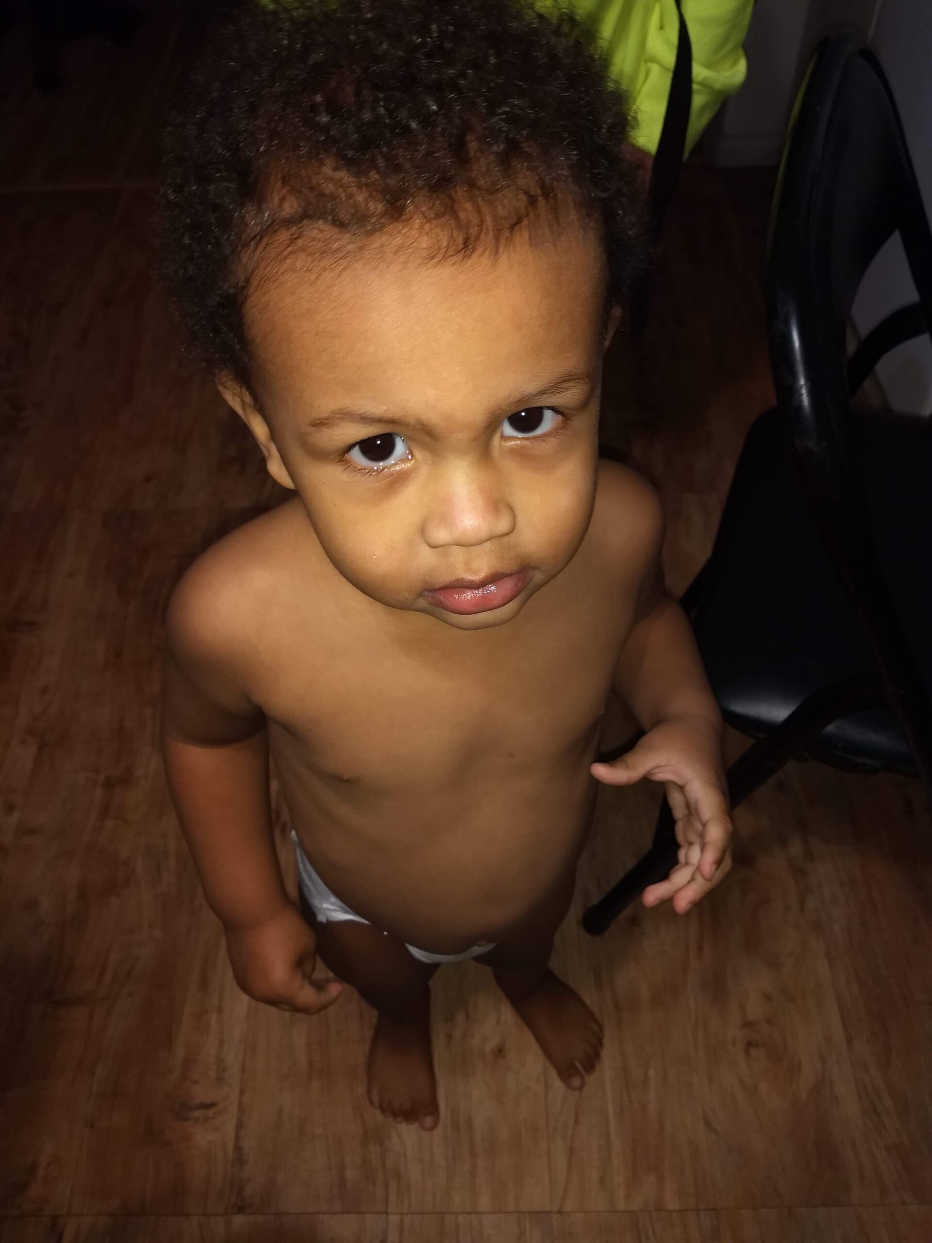 Young child standing barefoot on a wooden floor, shirtless and wearing a diaper, looking up at the camera with a serious expression.