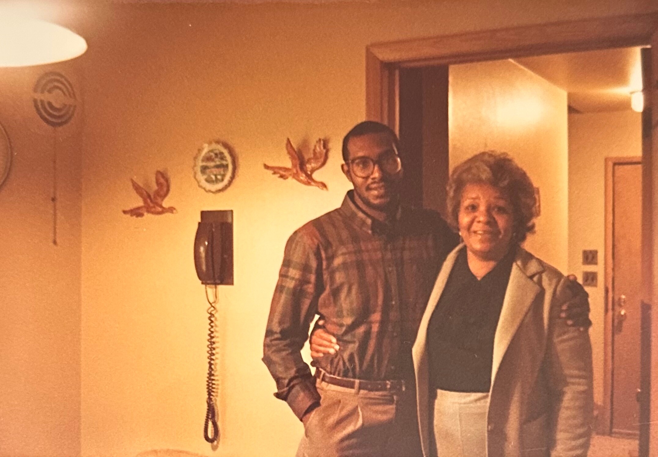 Two people pose together in a doorway inside a warmly lit home; a man in a plaid shirt and glasses stands beside an older woman smiling. Artwork and a wall phone are visible in the background.