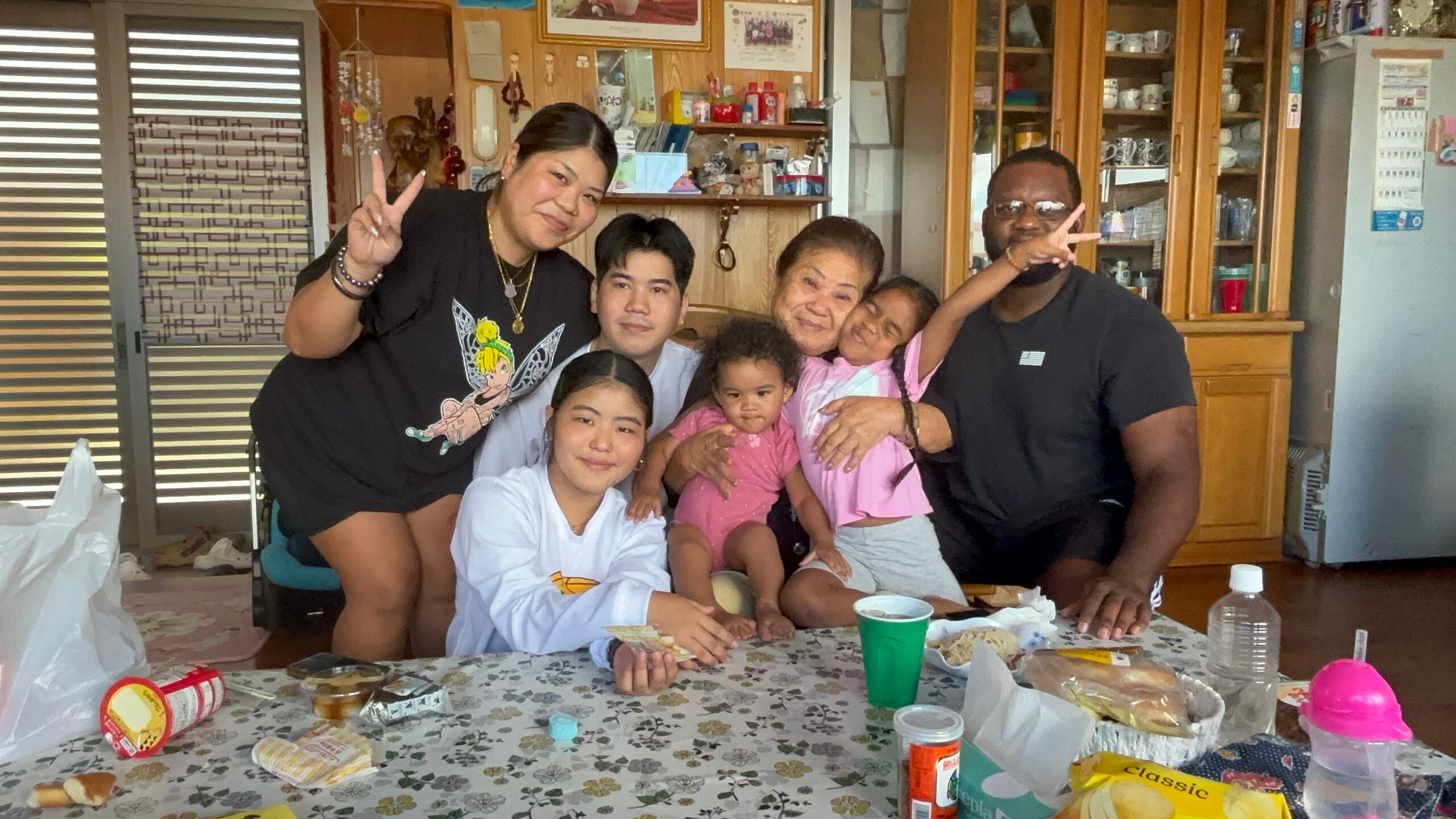 A large family group posing indoors, several people smiling and making peace signs around a table with snacks and cups.