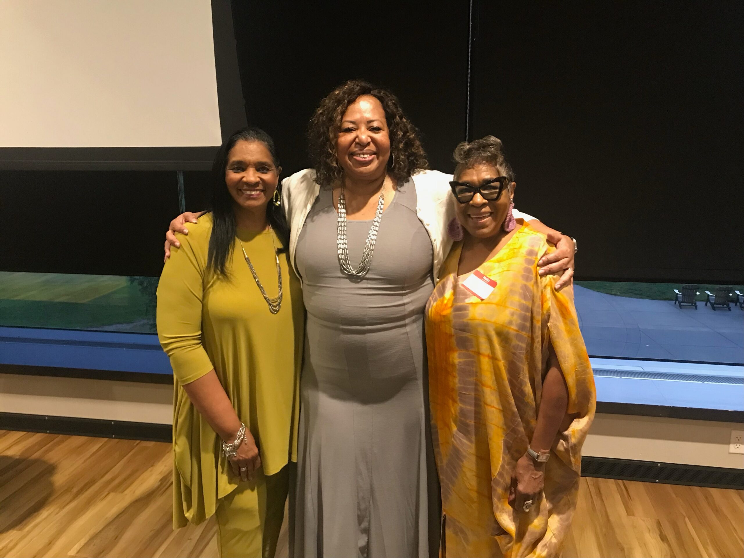 Three women posing together and smiling on a stage, wearing colorful dresses (yellow, grey, and orange) and accessorized with jewelry, arms around each other in a group photo.