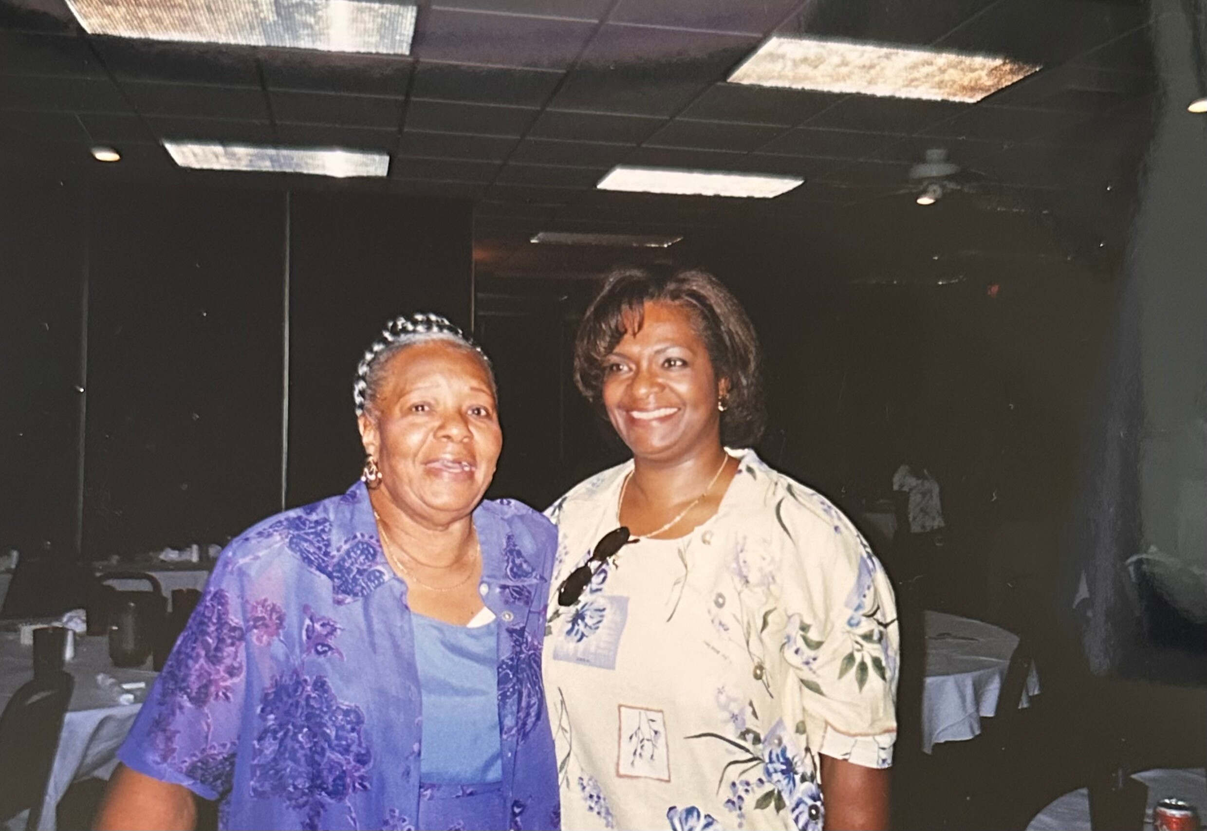Two women pose together and smile at a celebratory event, one in a purple floral top and the other in a light floral blouse with sunglasses hanging from her neckline.