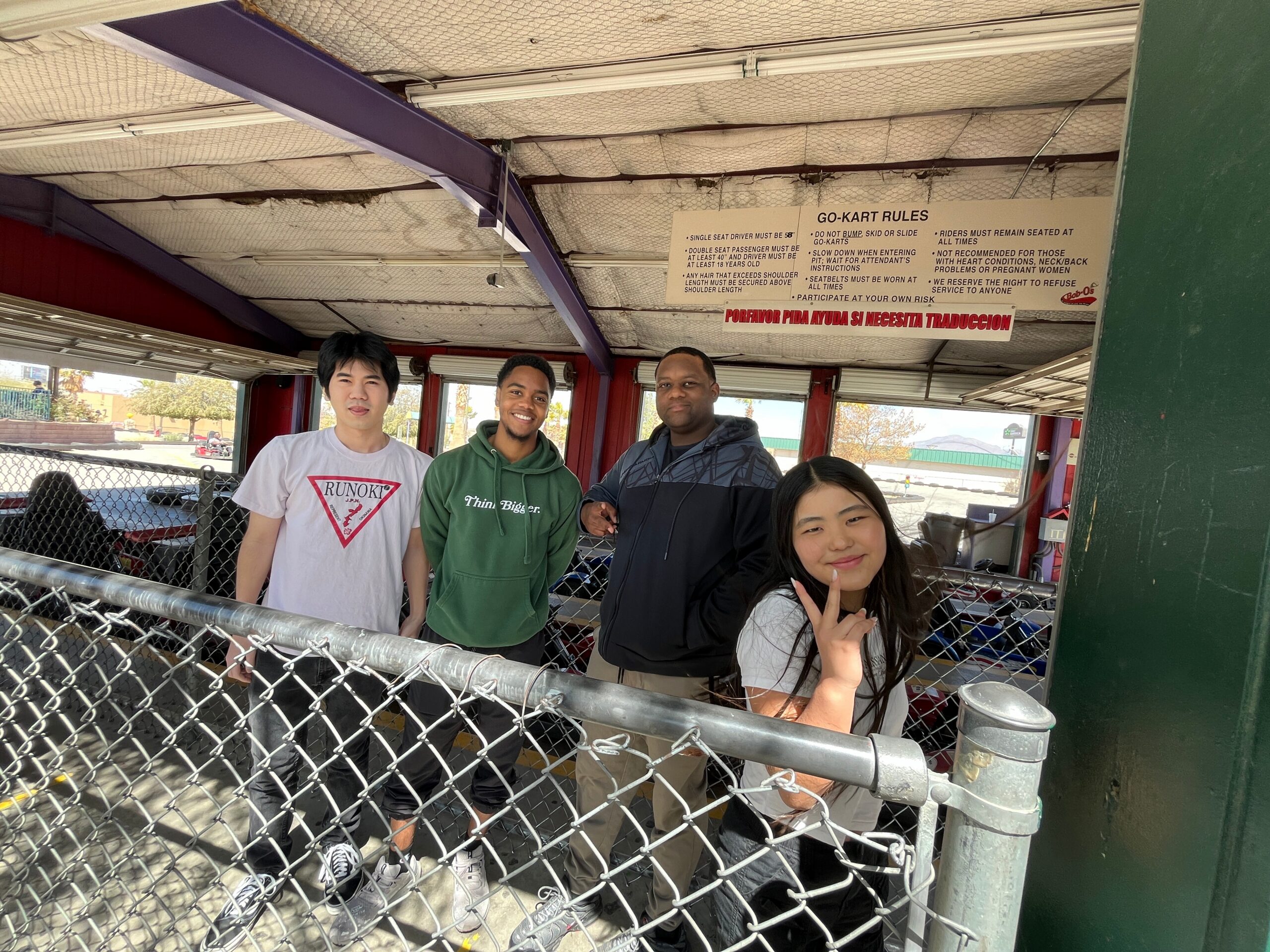 Four friends pose for a photo inside a go-kart queue area, with a chain-link fence in front and a GO-KART RULES sign visible above them.