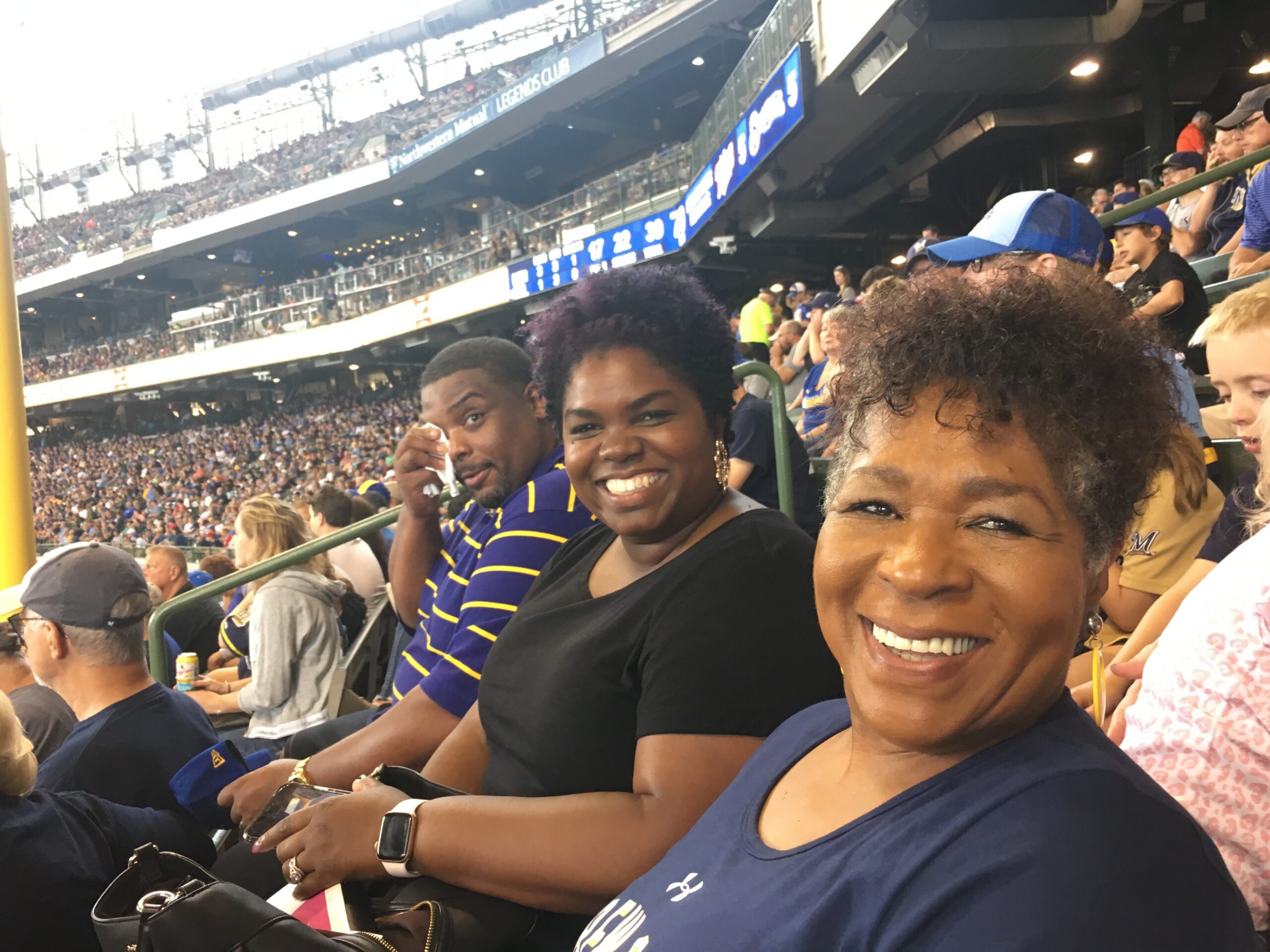 Smiling group of fans in blue at a stadium, cheering during a game with crowded stands in the background.