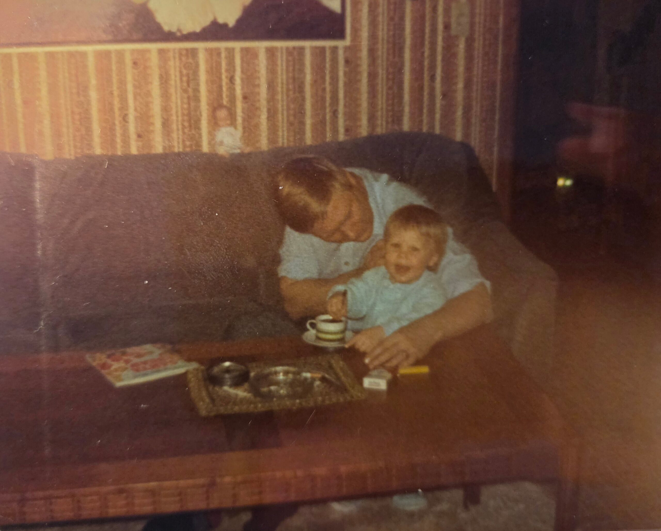 Two people sitting on a brown sofa in a living room with striped wallpaper, coffee table with a mug in front of them.
