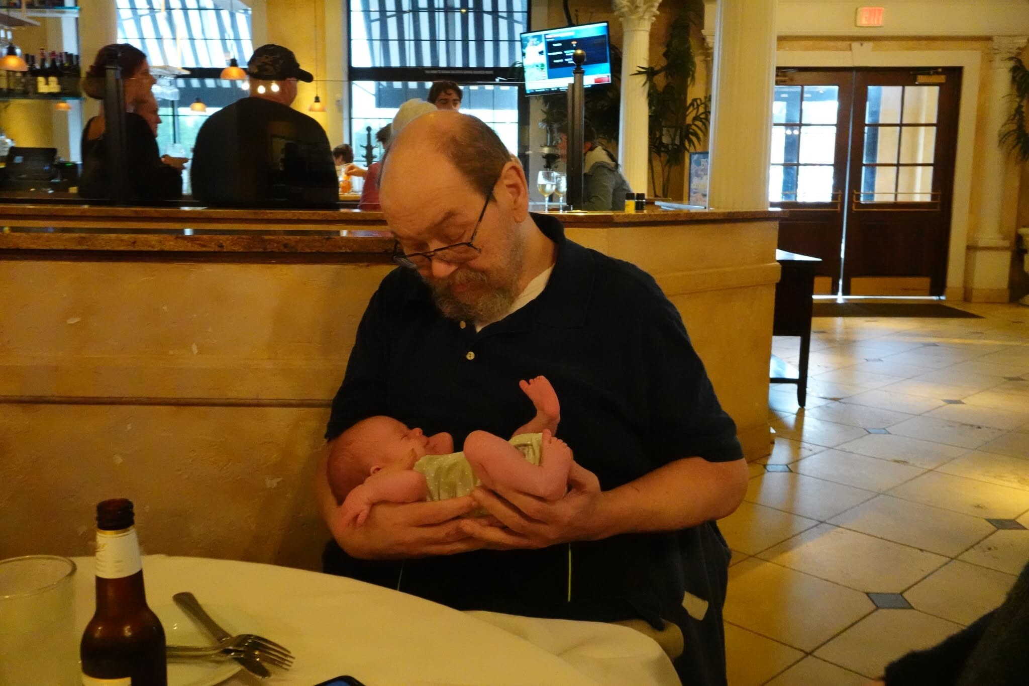 Bearded man with glasses cradling a newborn baby at a restaurant table.