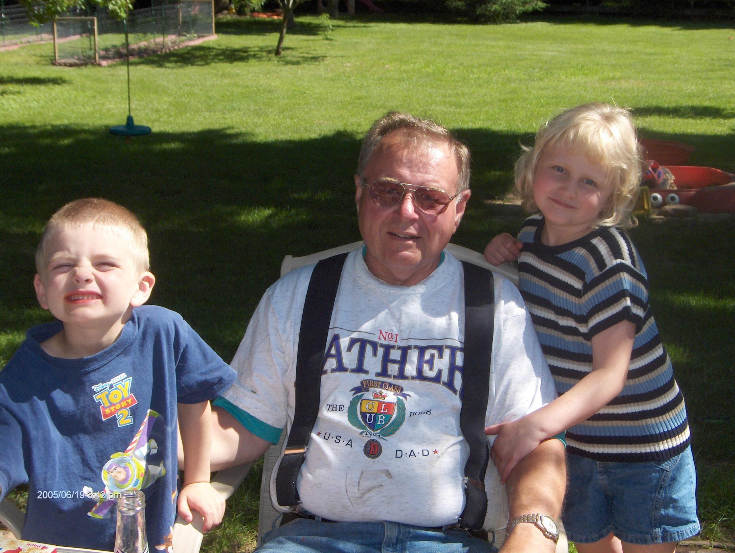 Smiling man wearing sunglasses poses with two children in a sunny grassy backyard.