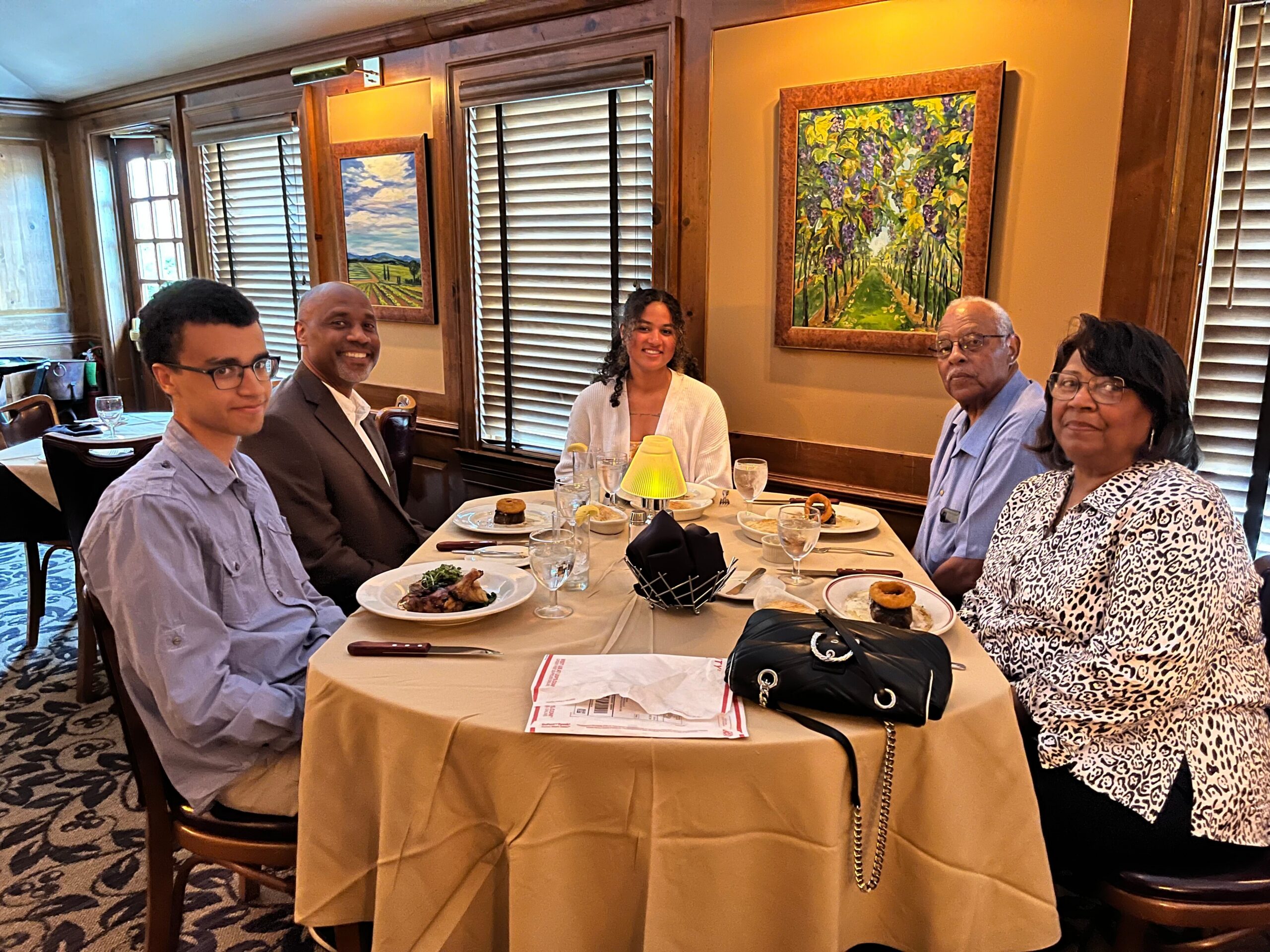 Group of five adults dining together at a restaurant table with plates, glasses, and a yellow napkin centerpiece in a wood-paneled room with framed paintings on the wall.