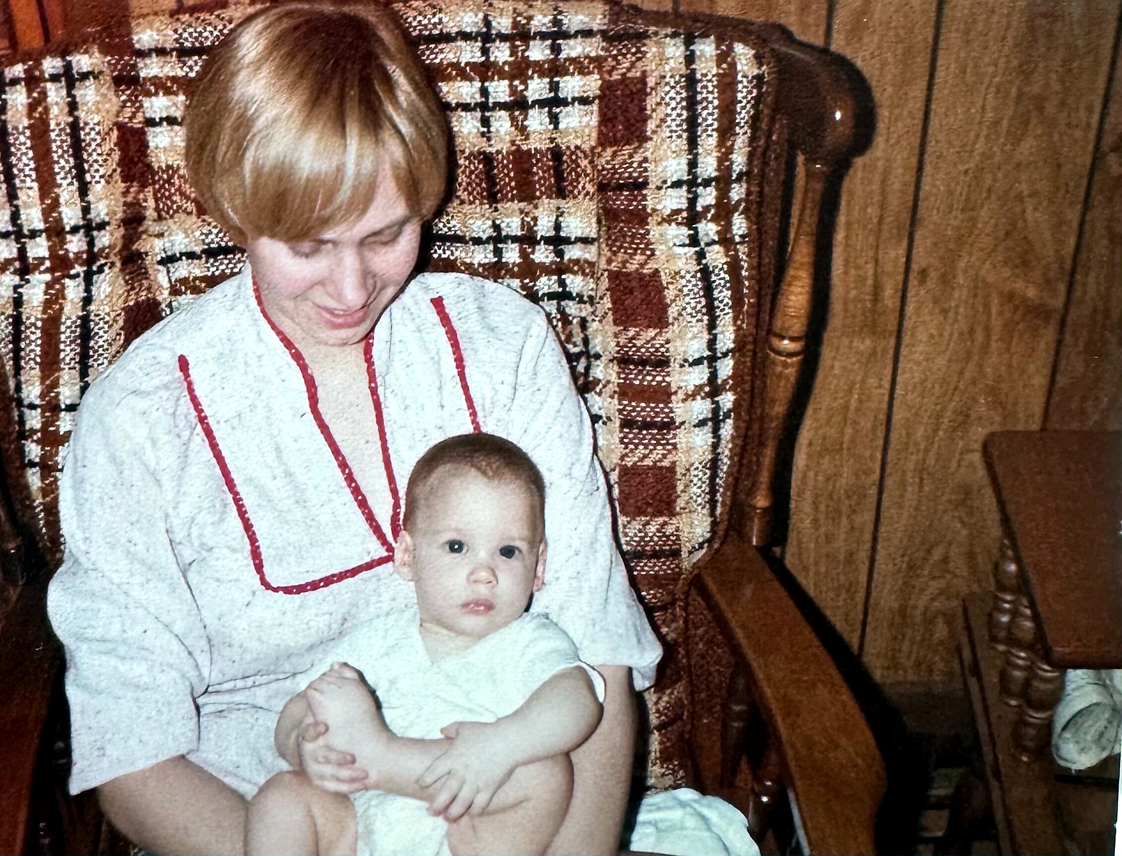Woman with short blonde hair sitting on a plaid couch while cradling a baby wrapped in a gray outfit with red trim.