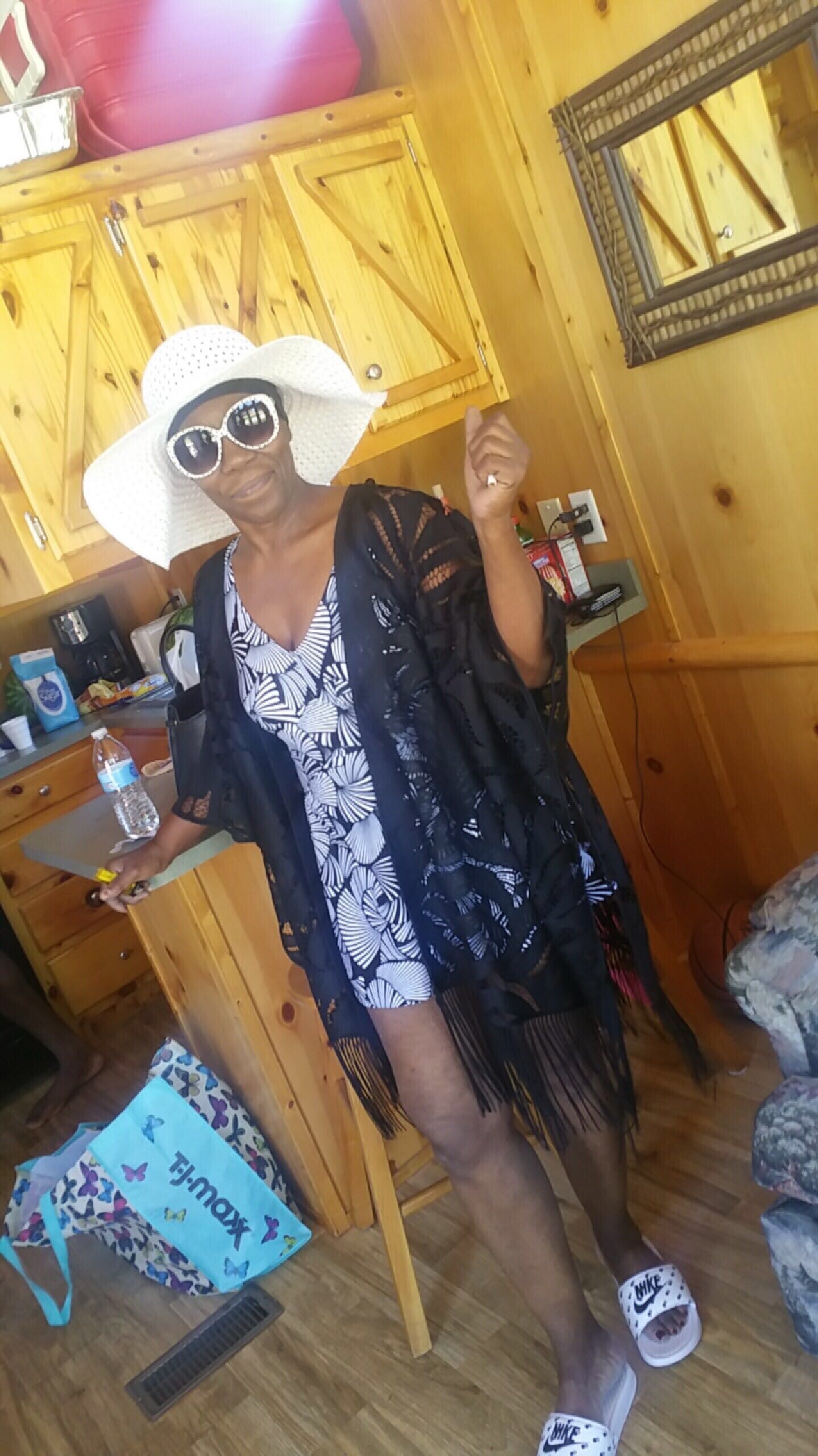 Woman in a wood-paneled kitchen wearing a large white sunhat and oversized white sunglasses, with a black fringed cover-up over a patterned dress, gesturing with her hand.