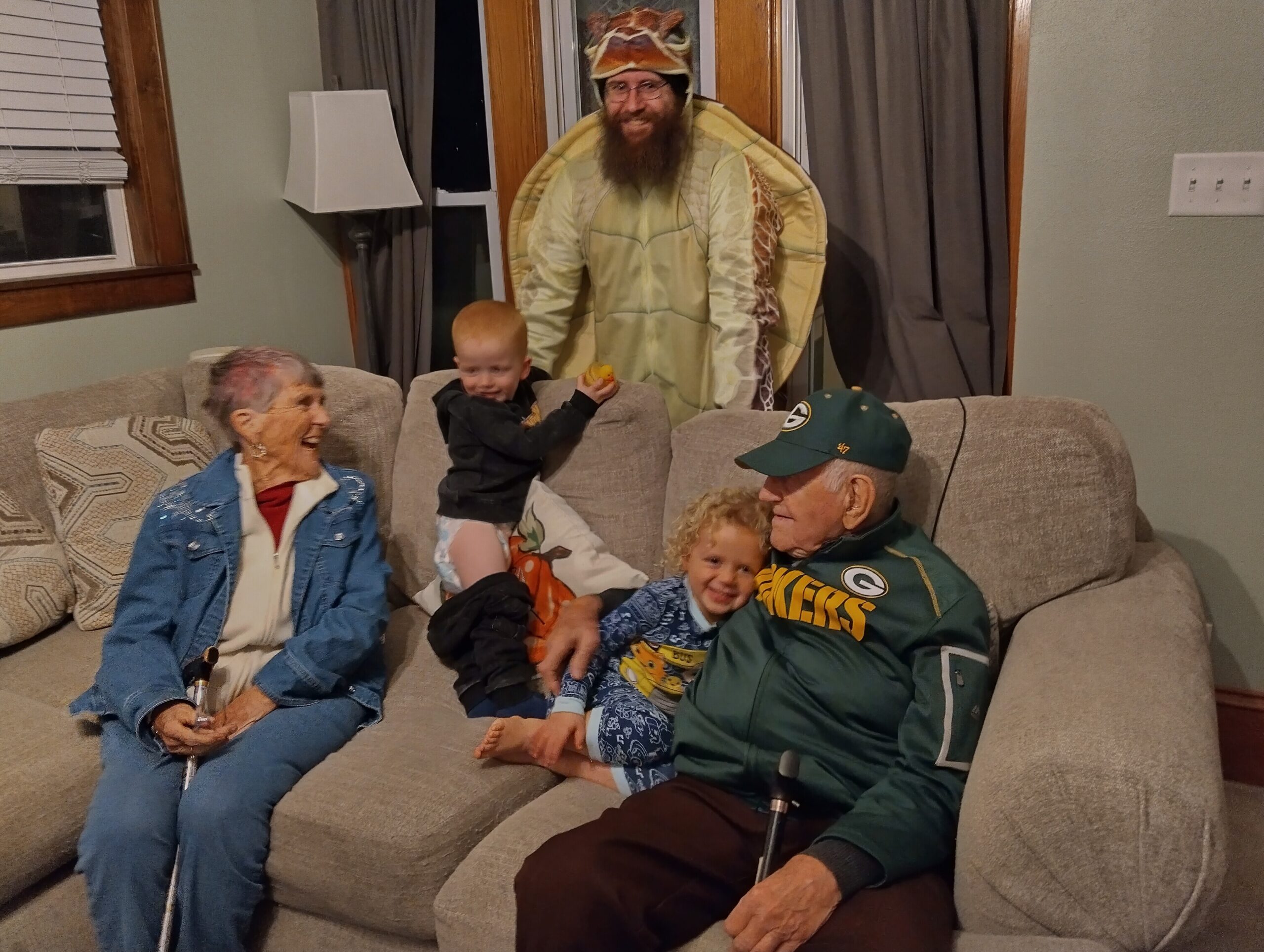 People in a living room wearing costumes on a couch; a child sits between an elderly woman and an adult man in a Packers jacket, while a man in a turtle suit looms behind them.