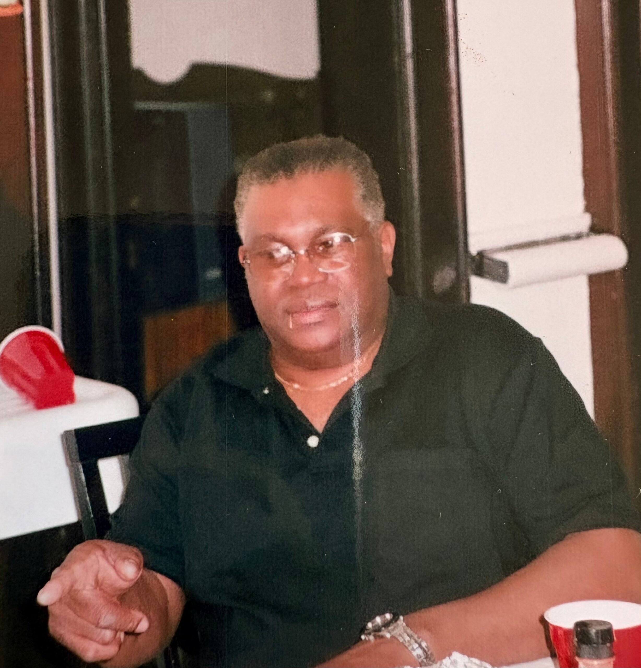 Older man with short gray hair and glasses sits at a table, clasping his hands, wearing a dark shirt; a red cup and small bottle are on the table behind him.
