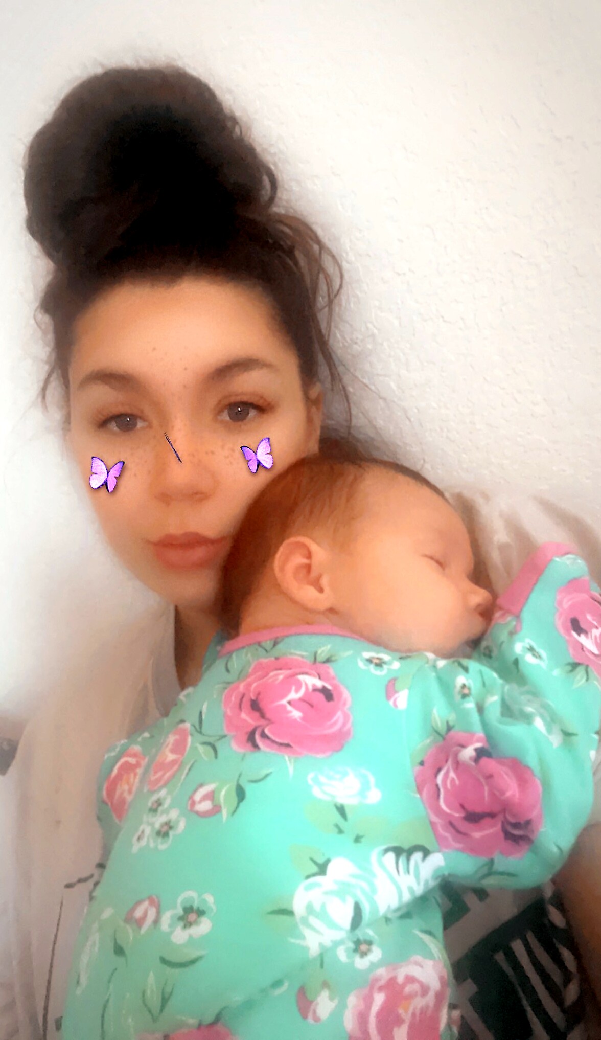 Woman with a dark hair bun cradling a sleeping baby against a light wall; baby is wrapped in a turquoise floral blanket.
