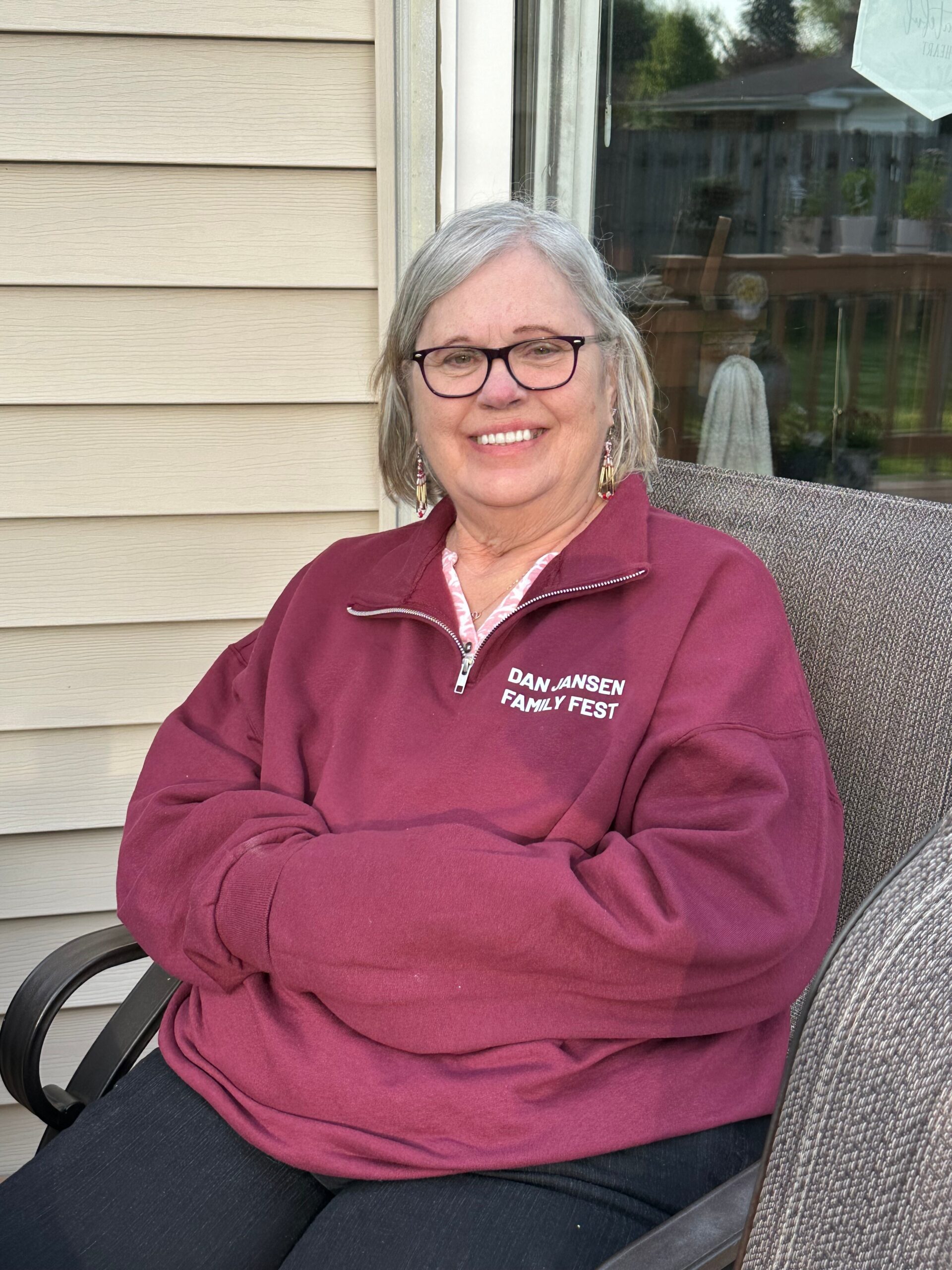 Smiling older woman with gray hair and glasses, wearing a maroon zip-up sweatshirt, seated on a patio chair outdoors with a house siding background.