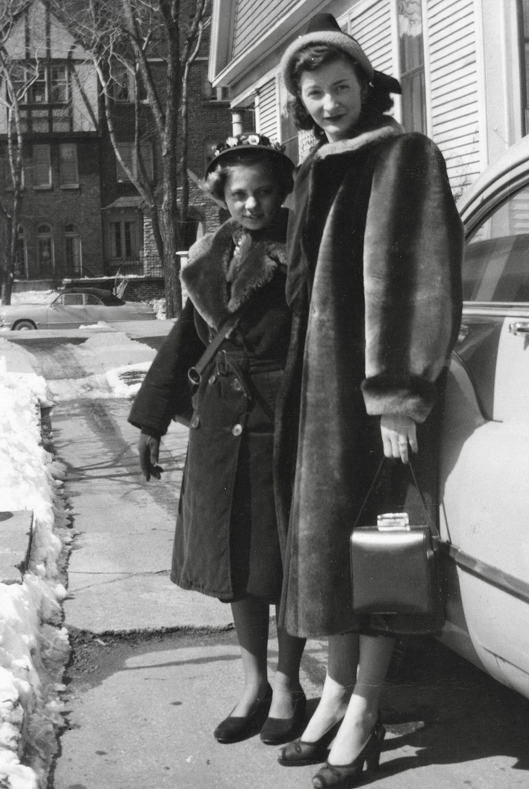 Two women in vintage fur coats and hats standing on a snowy sidewalk beside a car.