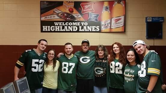 Eight people in Green Bay Packers jerseys pose together in a bowling alley under a 'Welcome to Highland Lanes' sign.