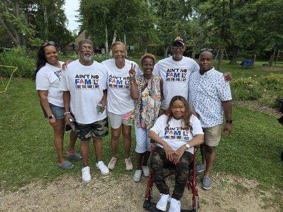 Group of eight adults posing in a park, all wearing matching shirts; one person sits in a wheelchair in front center.