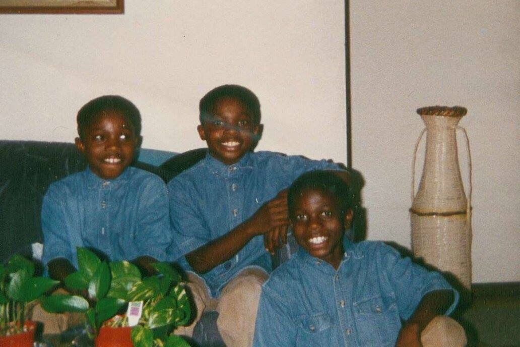 Three smiling Black boys wearing matching blue denim shirts sit on a couch indoors, with a potted plant on the left and a tall beige vase on the right.