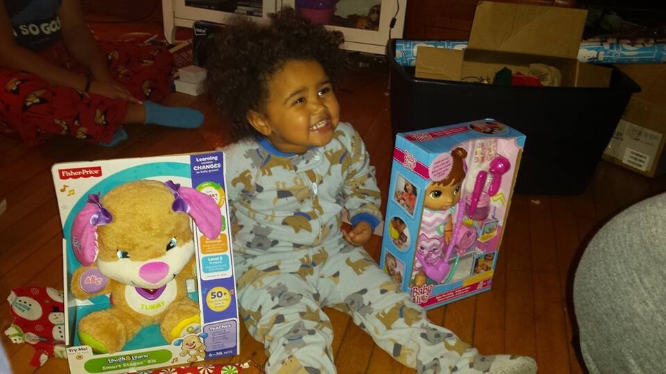 Smiling toddler in grey animal-print pajamas sits on a wooden floor, with two boxed toys nearby: a Fisher-Price plush puppy on the left and a pink doll playset on the right.