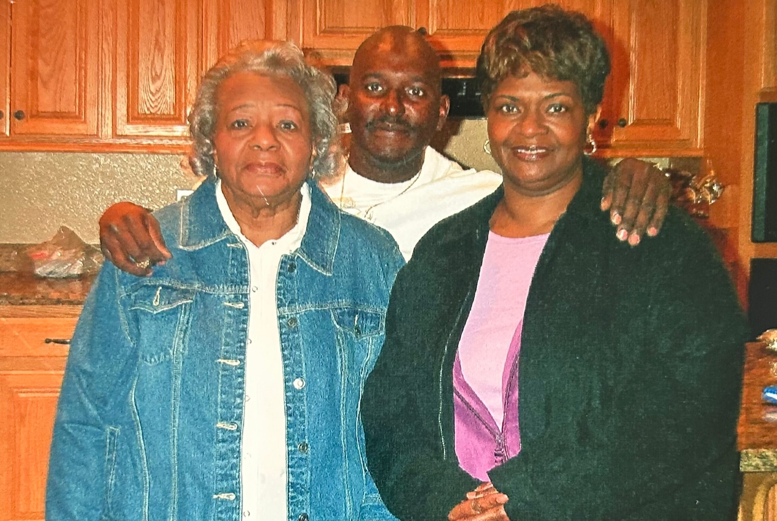 A family group posing in a kitchen: an elderly woman in a denim jacket, a man in the middle, and a smiling woman in a pink top on the right, all with arms around each other.