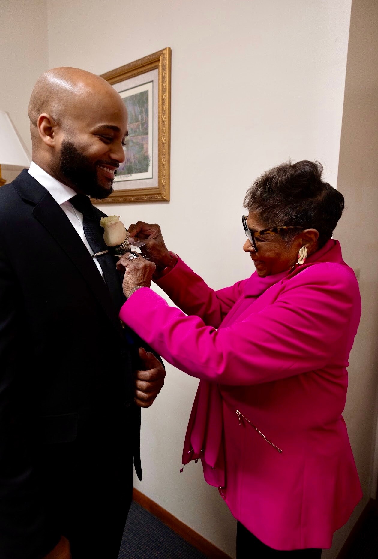 A man in a black suit smiles as a woman pins a white boutonniere to his lapel.