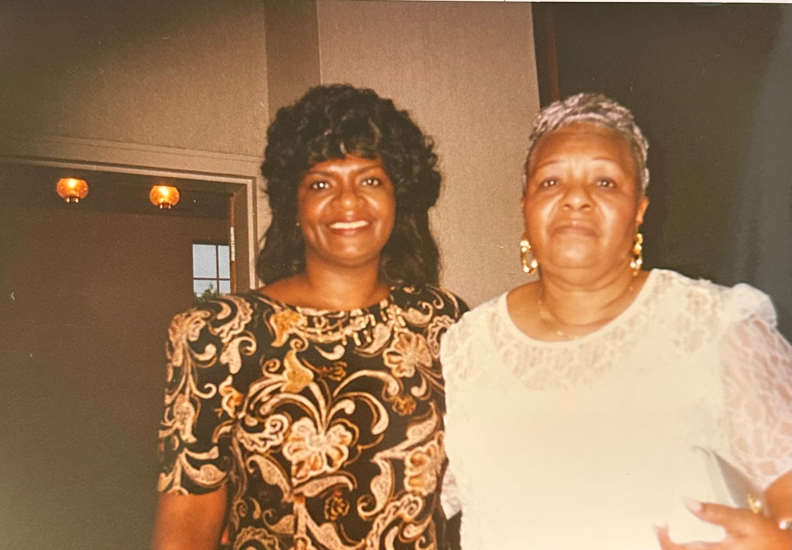 Two women pose together indoors, smiling, one in a black-and-gold patterned dress and the other in a light blouse with short gray hair.