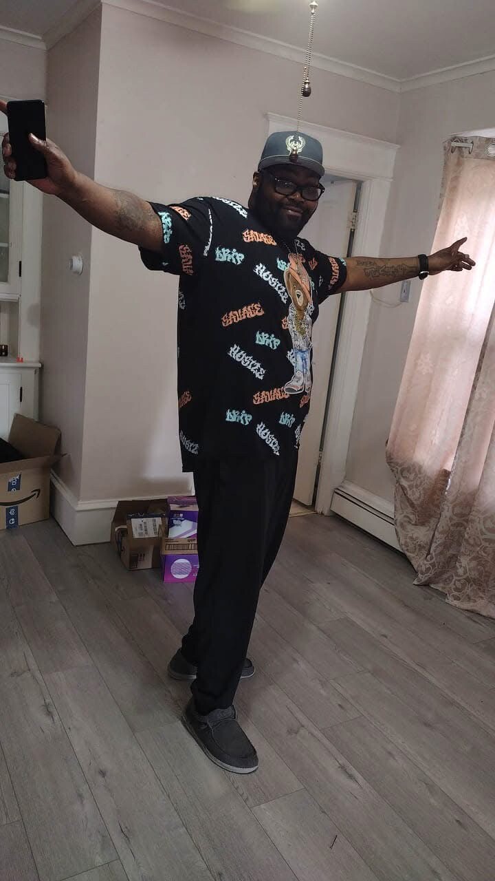 Man indoors posing with arms outstretched, wearing a black graphic T-shirt, cap, glasses, and gray sneakers in a living room-like space.