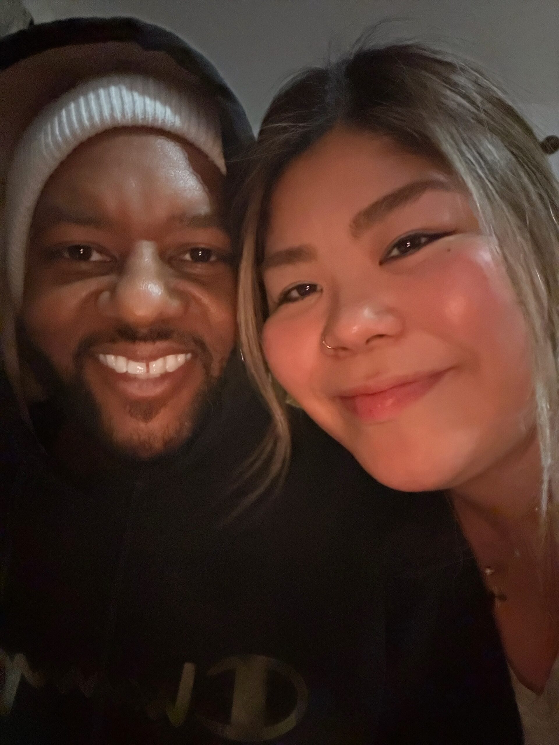 Close-up selfie of two people smiling, a man in a hoodie wearing a white knit cap and a woman with light hair leaning in close.