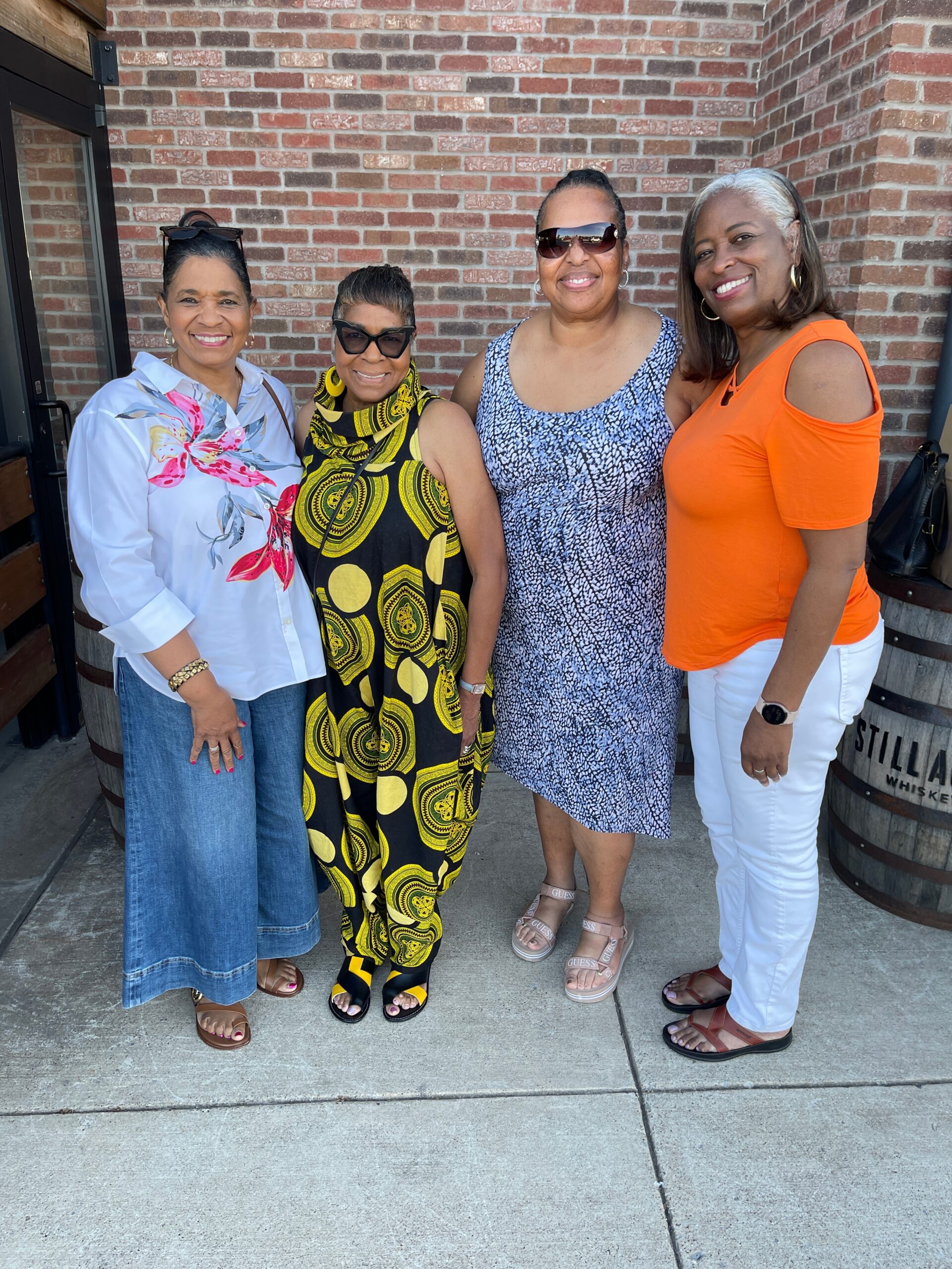 Four women standing close together outdoors in front of a brick wall, smiling at the camera, wearing colorful summer outfits.