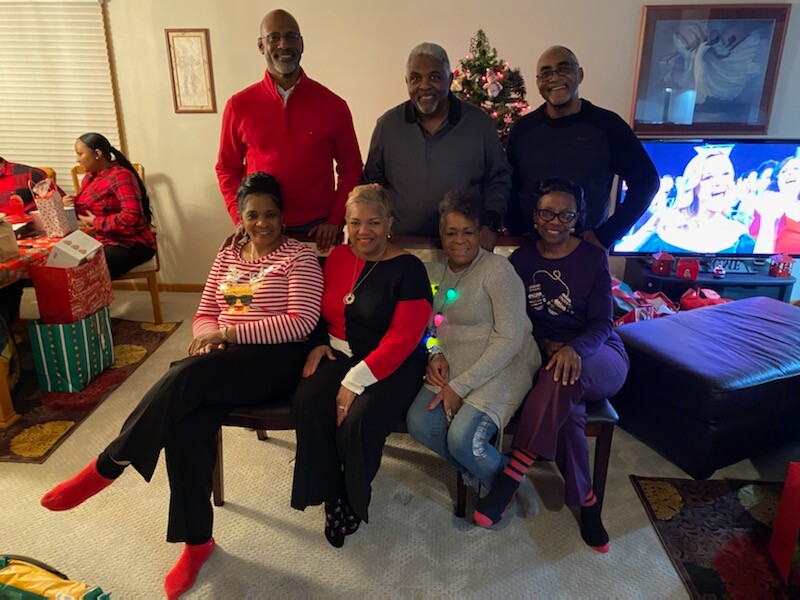 Eight adults posing for a Christmas photo in a living room, with a Christmas tree and presents nearby.