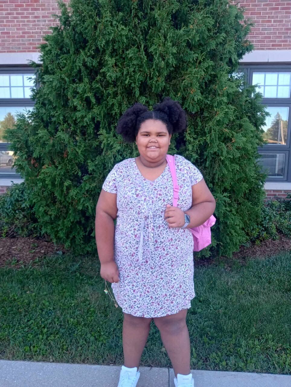 Girl with two Afro puffs wearing a floral dress and white sneakers, standing in front of a large evergreen shrub with a pink backpack beside her.