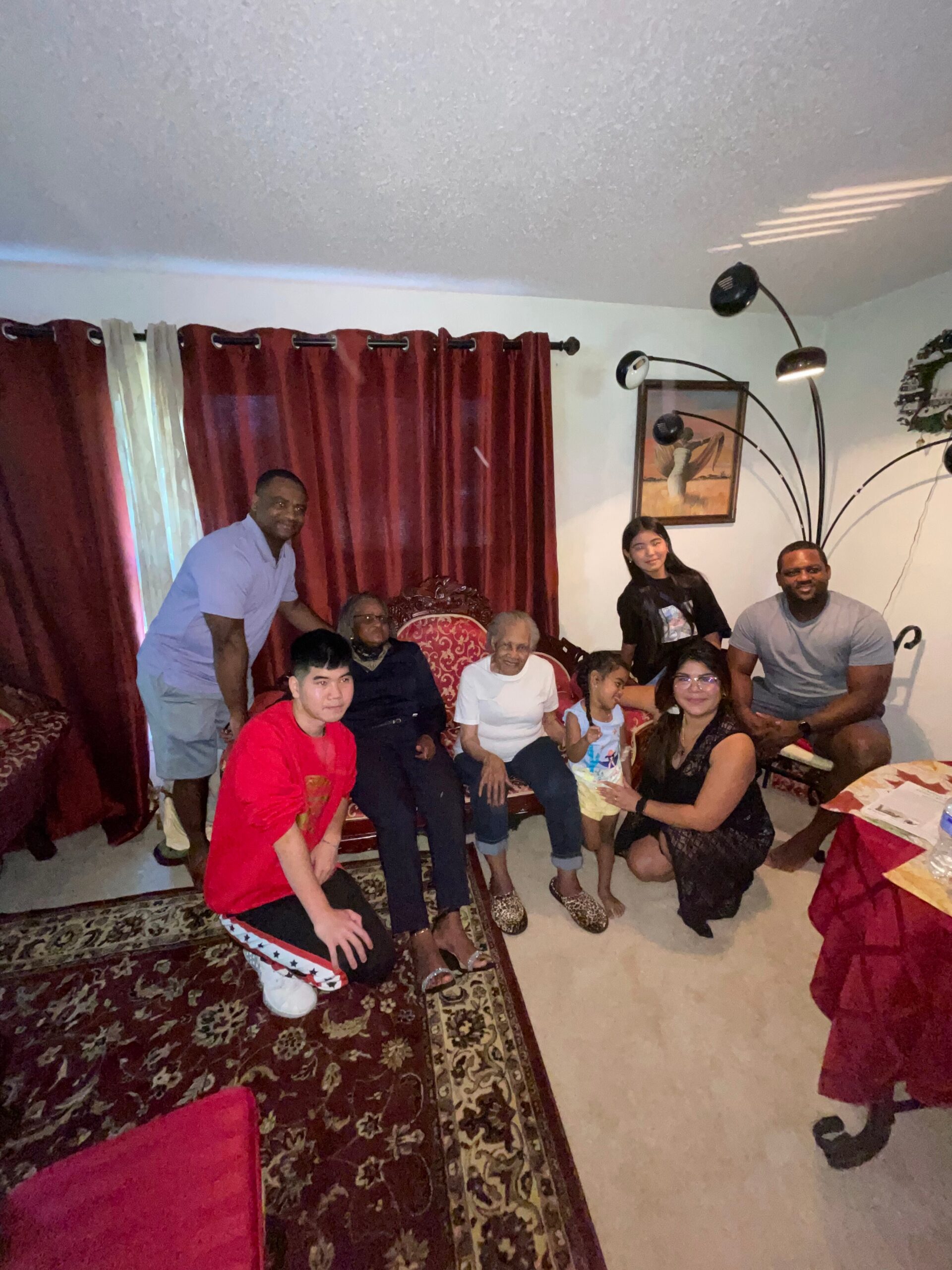 Family group posing for a photo in a living room with red curtains, a patterned rug, and several adults and children gathered together.