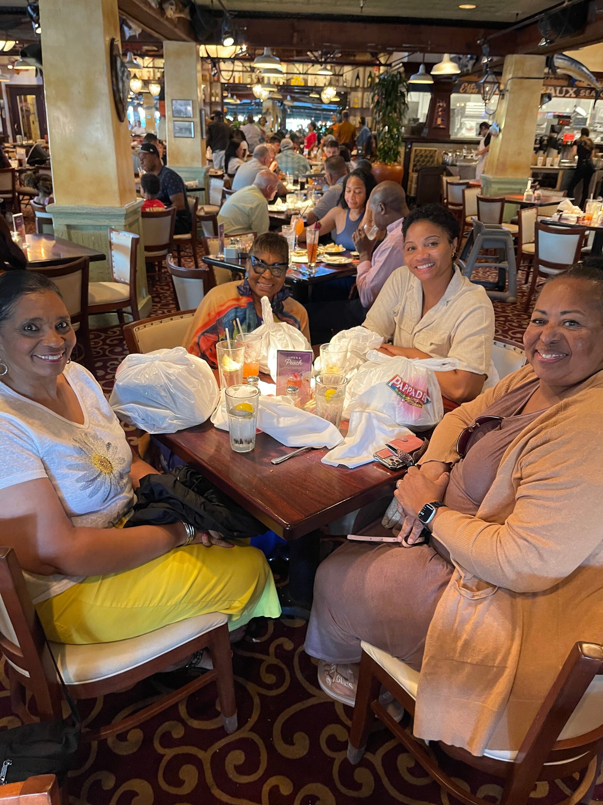 Group of four women smiling and sitting around a restaurant table with drinks and takeaway bags, enjoying a meal together.