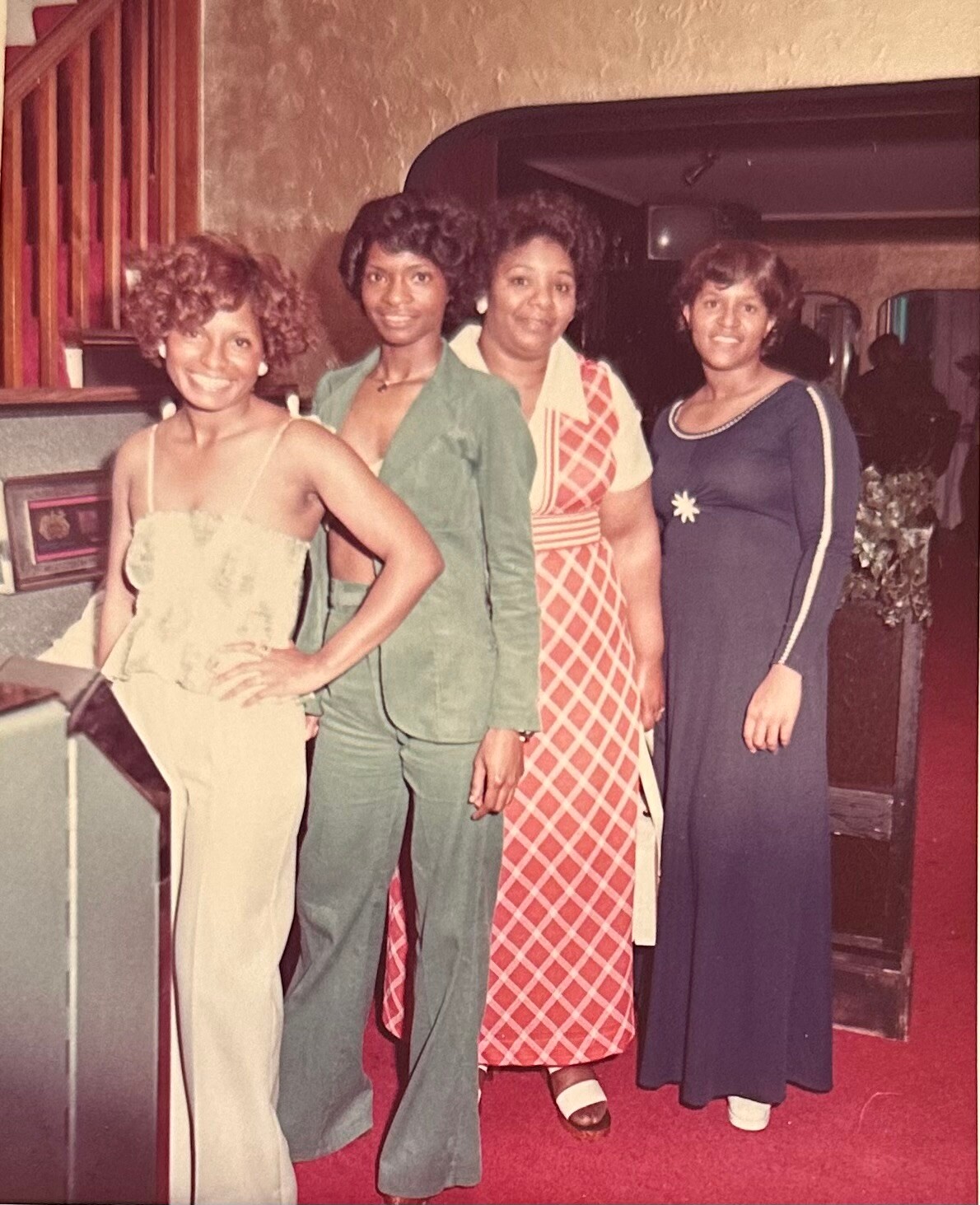 Four women in 1970s attire posing together at a social event, standing in a line and smiling at the camera.