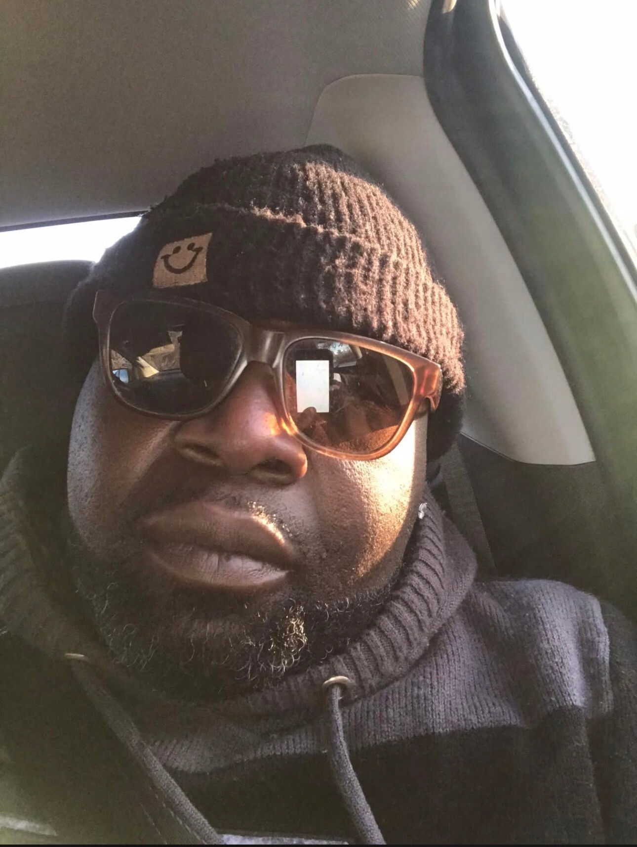 Close-up of a man in a brown knit beanie and brown sunglasses inside a car, wearing a dark hoodie.