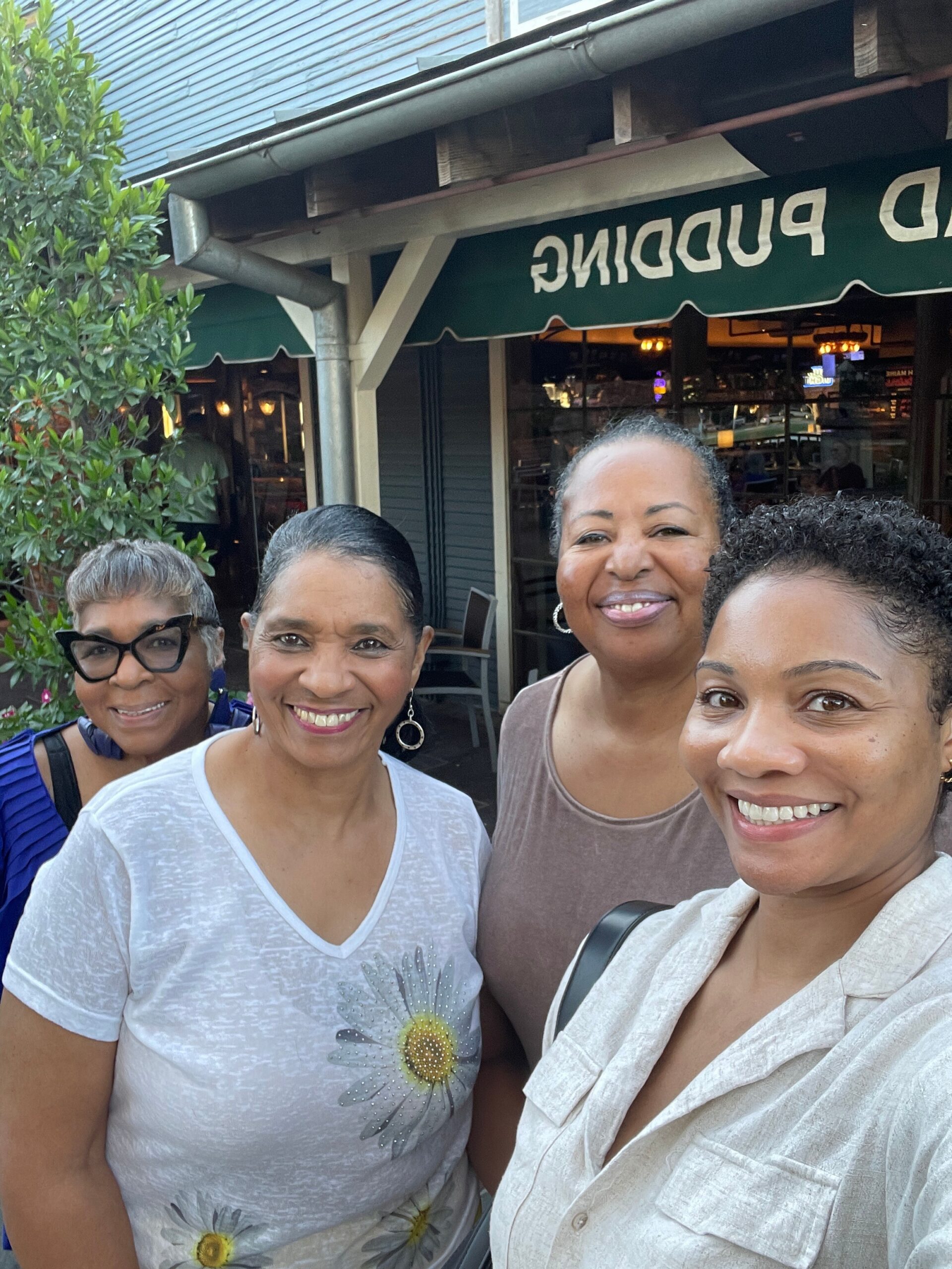 Four women posing for a group selfie outside a cafe/restaurant with a green awning. All are smiling and close together.
