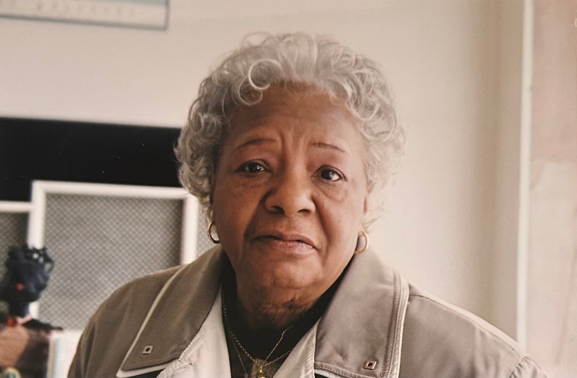 Portrait of an elderly woman with short gray curly hair, wearing a beige jacket, indoors near a camera setup.