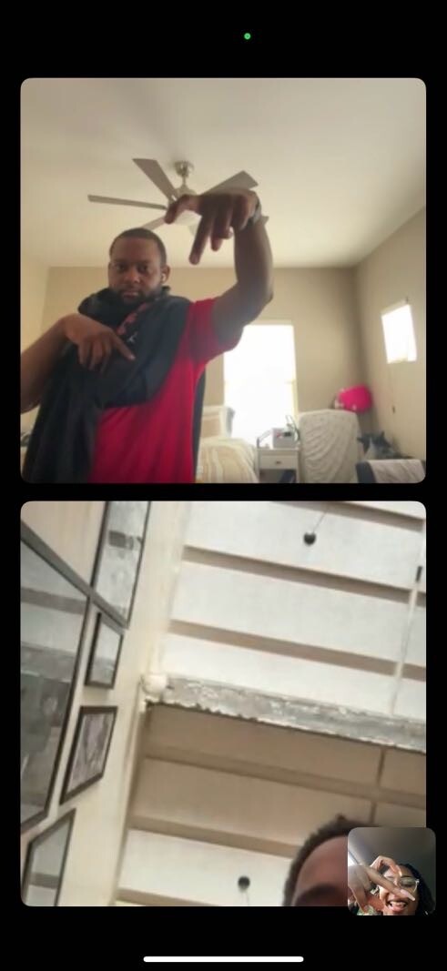 Man in a red shirt holding a jacket and pointing toward the camera in a sunlit room (video call).