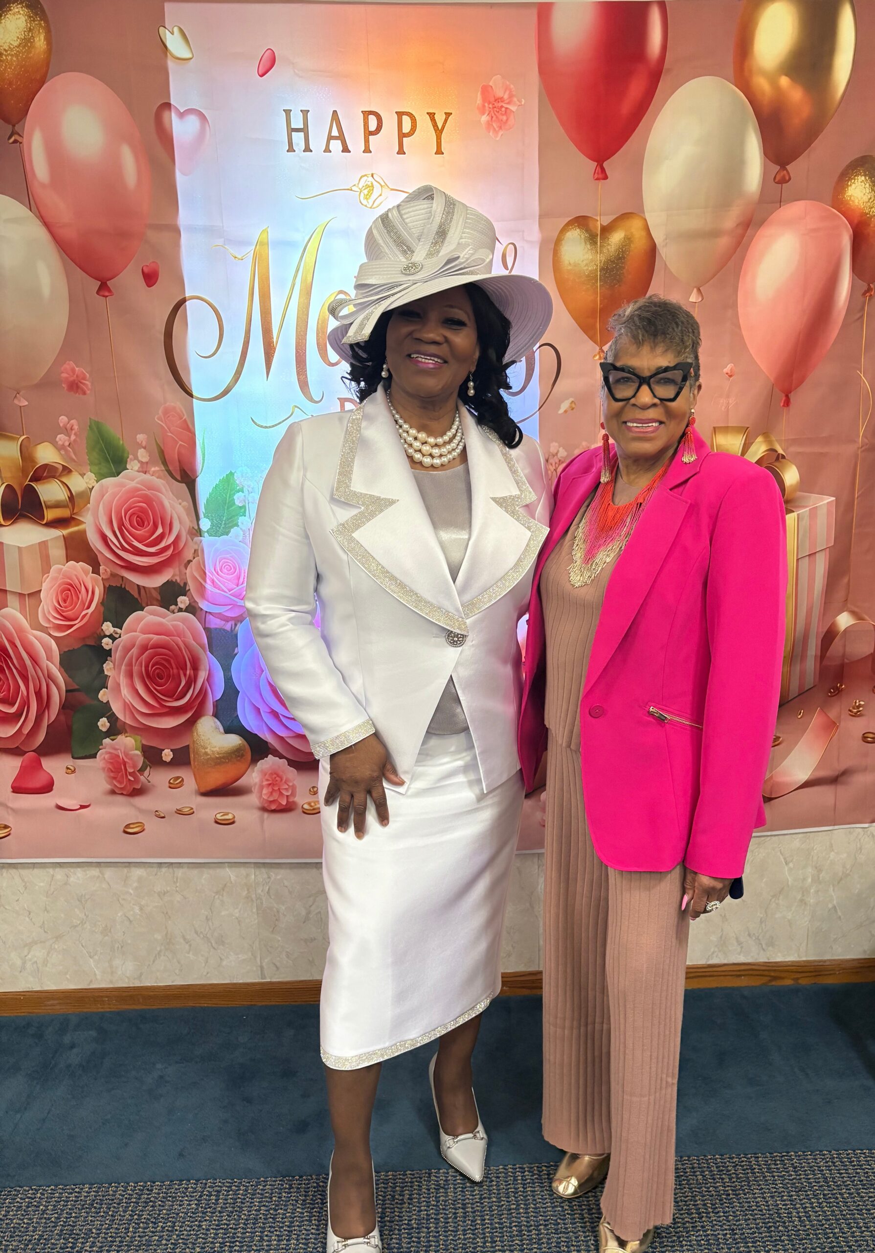 Two women in elegant outfits pose together at a celebration backdrop with balloons and roses behind them.