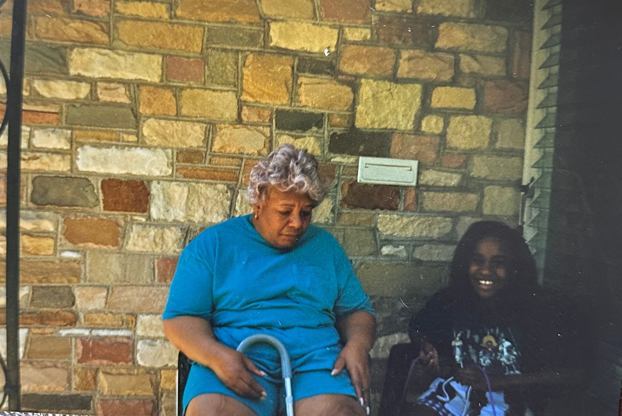 Two women sit together against a stone exterior wall; the woman on the left wears a bright blue shirt and uses a walker, while another person sits to her right smiling.