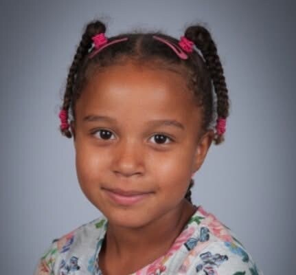 Portrait of a smiling young girl with braided pigtails tied with pink bands, wearing a floral shirt.
