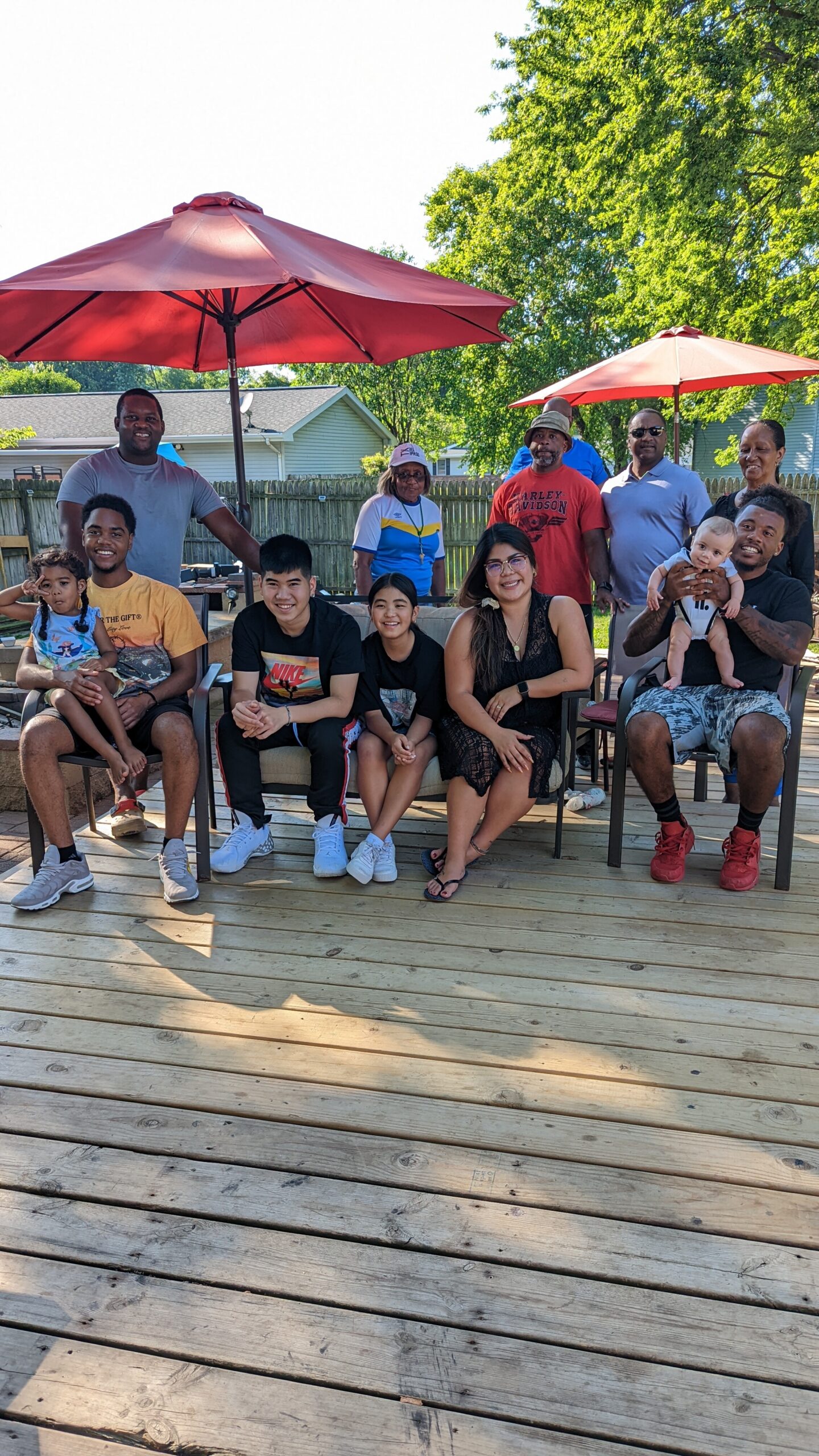 Group of diverse family members posing together on a wooden deck under red patio umbrellas in a sunny backyard.
