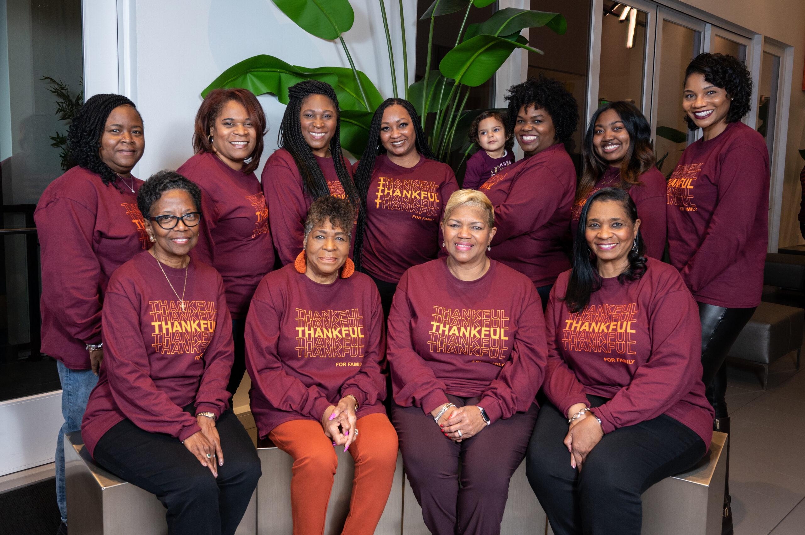 Group of diverse women (and a small child) wearing maroon shirts with 'THANKFUL' text, posing together in a bright indoor space.