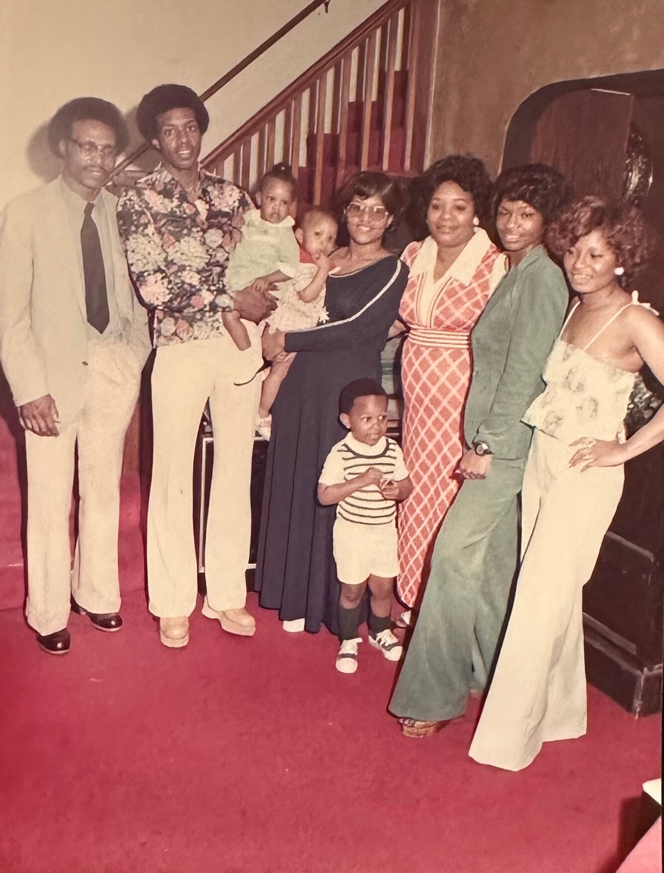 Family poses for a group photo on a red carpet indoors, with a staircase in the background and two small children in front.